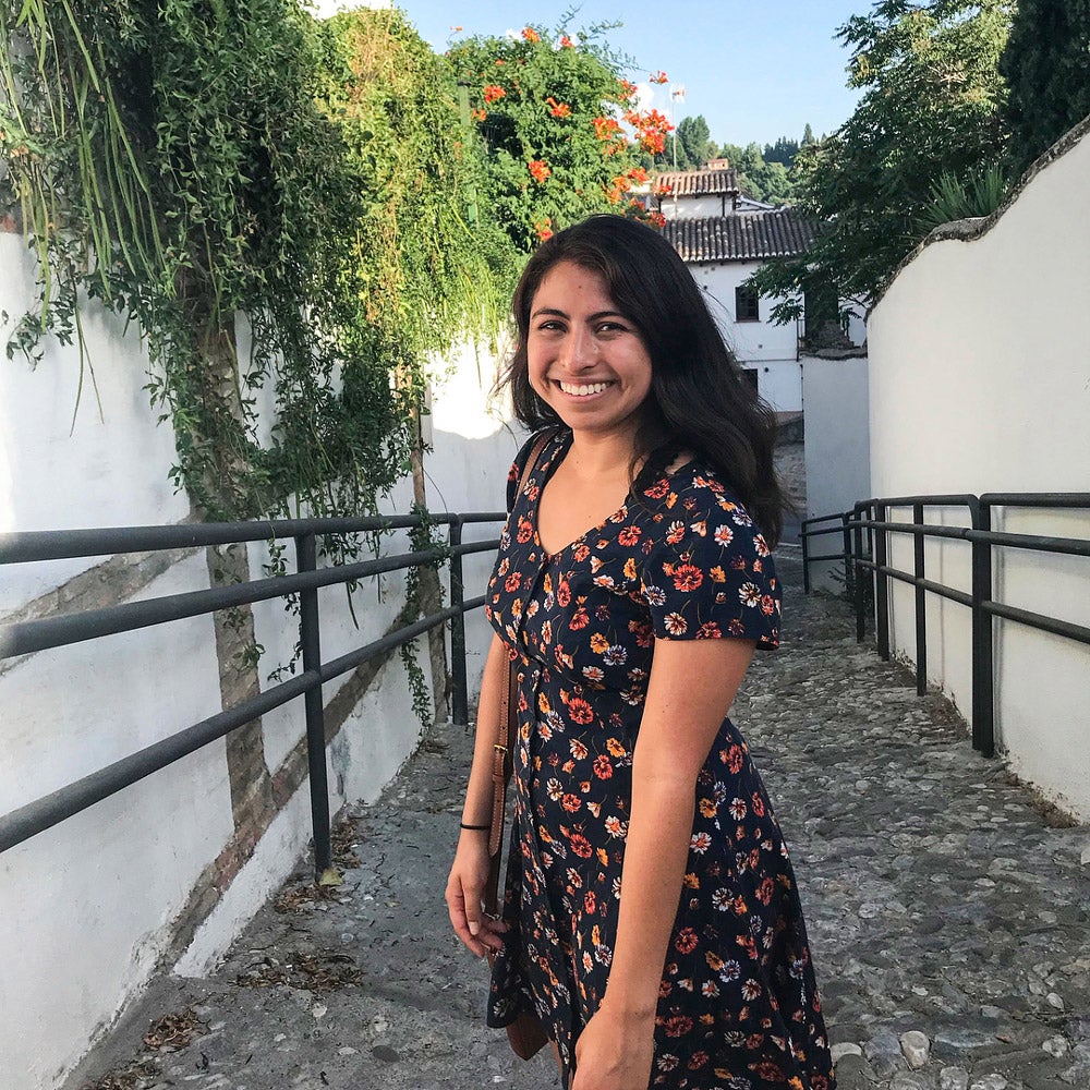 Smiling student wearing a floral dress standing atop a stone-paved walkway.