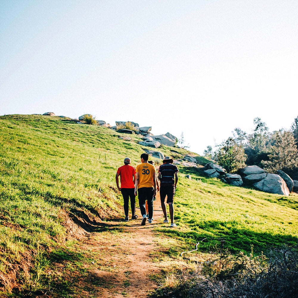 Three UCR students taking a hike along a dirt trail that is surrounded by large rocks and green grass.