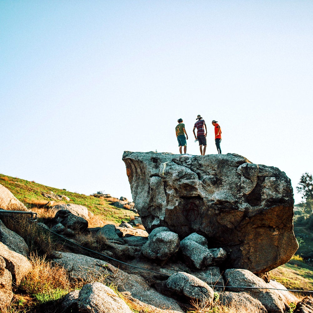 Three UCR students standing atop a giant boulder enjoying the view of blue sky and green grass.
