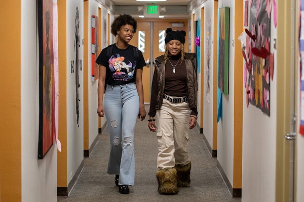 Two female students are walking down a narrow hall together while smiling.