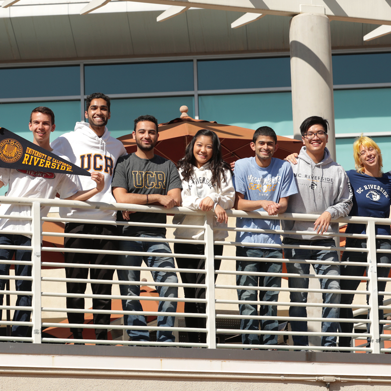 A group of students pose in a line outside at the Highlander Union Building Plaza on the UCR campus. They are leaning against a railing and are wearing UCR T-shirts.