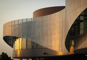 A view of the second level exterior of UCR's Student Recreation Center. The sun is setting providing an orange glow across the curves of the modern building.