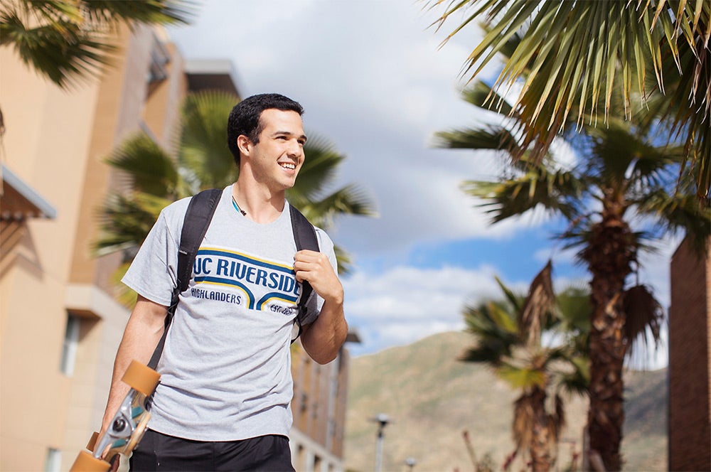 A male student walks through a sidewalk path of the UCR residence halls. He is carrying a skateboard in his right hand, and has his left hand on his backpack strap by his shoulder. The Student wears a UC Riverside T-shirt. There are palm trees lining the walkway and the background has a building, mountains and blue skies with clouds.