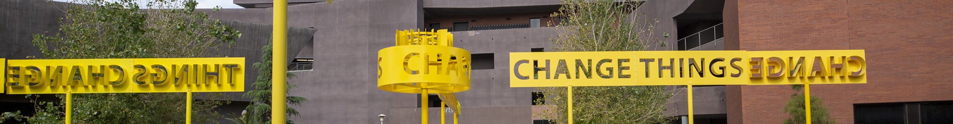 A bright yellow outdoor art installation near the Arts building on the UC Riverside campus featuring the words "CHANGE THINGS" in large, cutout letters against a backdrop of modern university buildings. The text is displayed both forward-facing and mirrored on elevated metal banners and a circular structure.