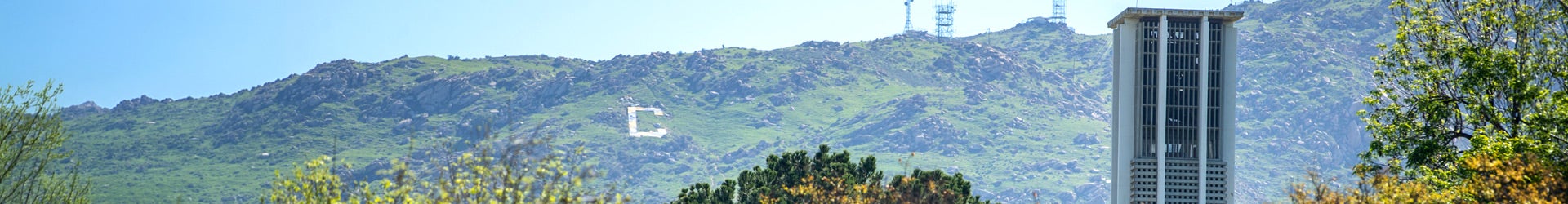 A wide landscape view of the University of California, Riverside campus, featuring the iconic concrete Bell Tower in the foreground. In the background, a large white "C" is visible on the green, rocky slopes of the Box Springs Mountains.