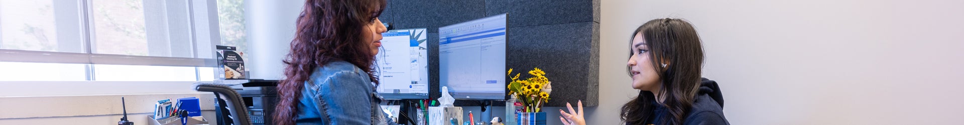 A UCR counselor with long, dark hair advises a female student in an office with dual computer monitors and sunflowers.
