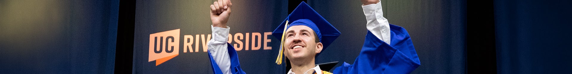 A happy male UCR graduate in a blue cap and gown raises his arms in celebration in front of a UC Riverside logo.