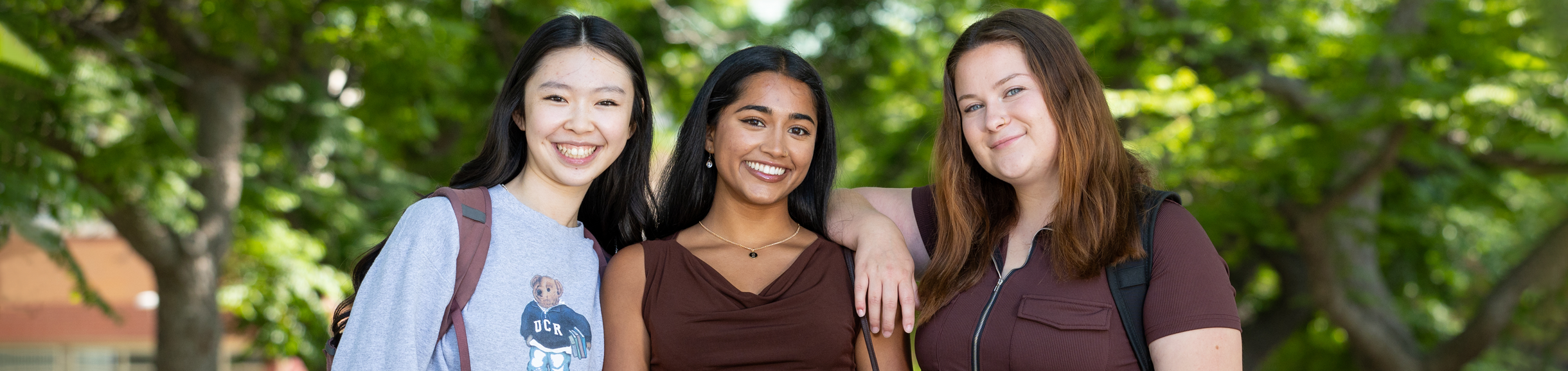 Three female students pose for a group photo on the UC Riverside campus. They are standing beneath trees that have bright green leaves on them.