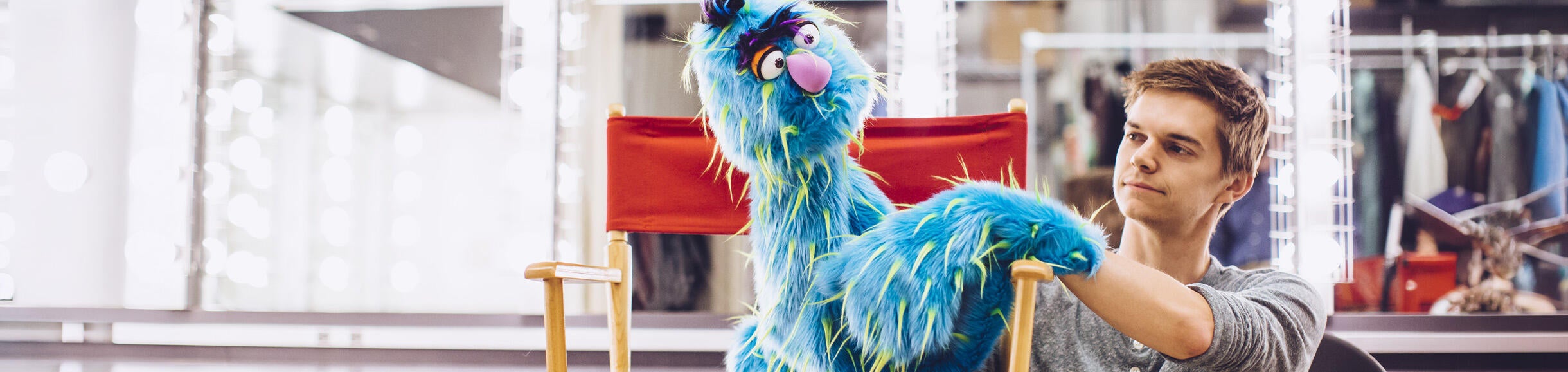 A man adjusts a shaggy blue monster puppet. The background features mirrors and clothing racks, suggesting a theater setting.
