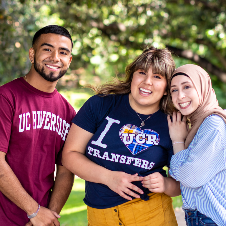 Three UC Riverside students pose for a mini group photo. A female student is flanked by a male student on the left side and another female student on the right side of the image. The male student is wearing a maroon colored T-shirt. The student in the middle is wearing a "I Heart Transfers" with the UCR letters inside the heart. While the third student is wearing a pinstriped shirt and a Hijab.