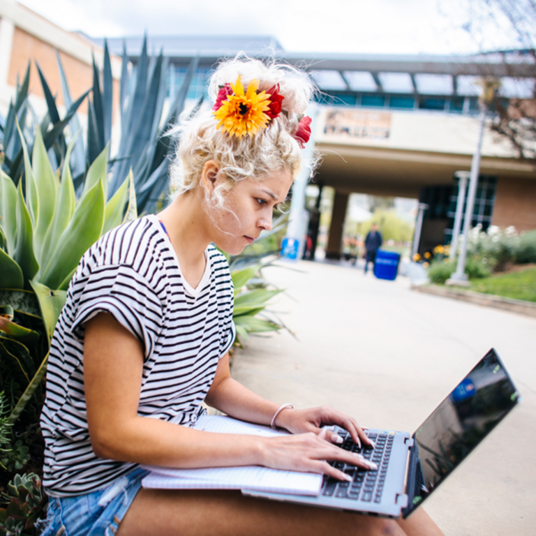 A female student types away at her laptop. She is sitting on the ground next to succulent plants outside Costo Hall on the UC Riverside campus.