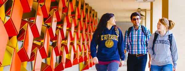 Two women in UC Riverside sweatshirts and a man in plaid shirt are talking as they walk past a geometrical art installation on the side of the Genomics building at UCR