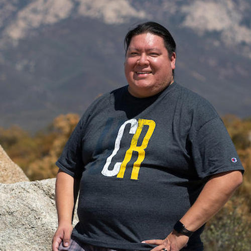 Doctoral student William Madrigal Jr., a member of the Cahuilla Band of Indians, smiling, stands in the desert. 