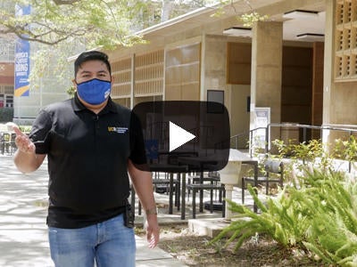 A male student, wearing a protective mask and black polo, leads students on a virtual campus tour.