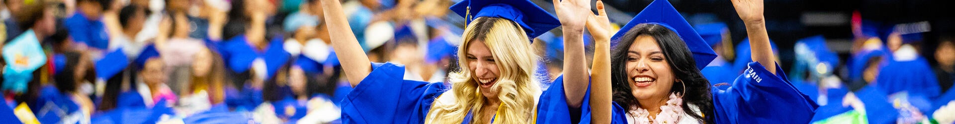 Two female students celebrate with their hands up while wearing blue caps and gowns at a UC Riverside commencement ceremony held in Ontario, Calif., at the end of spring quarter.