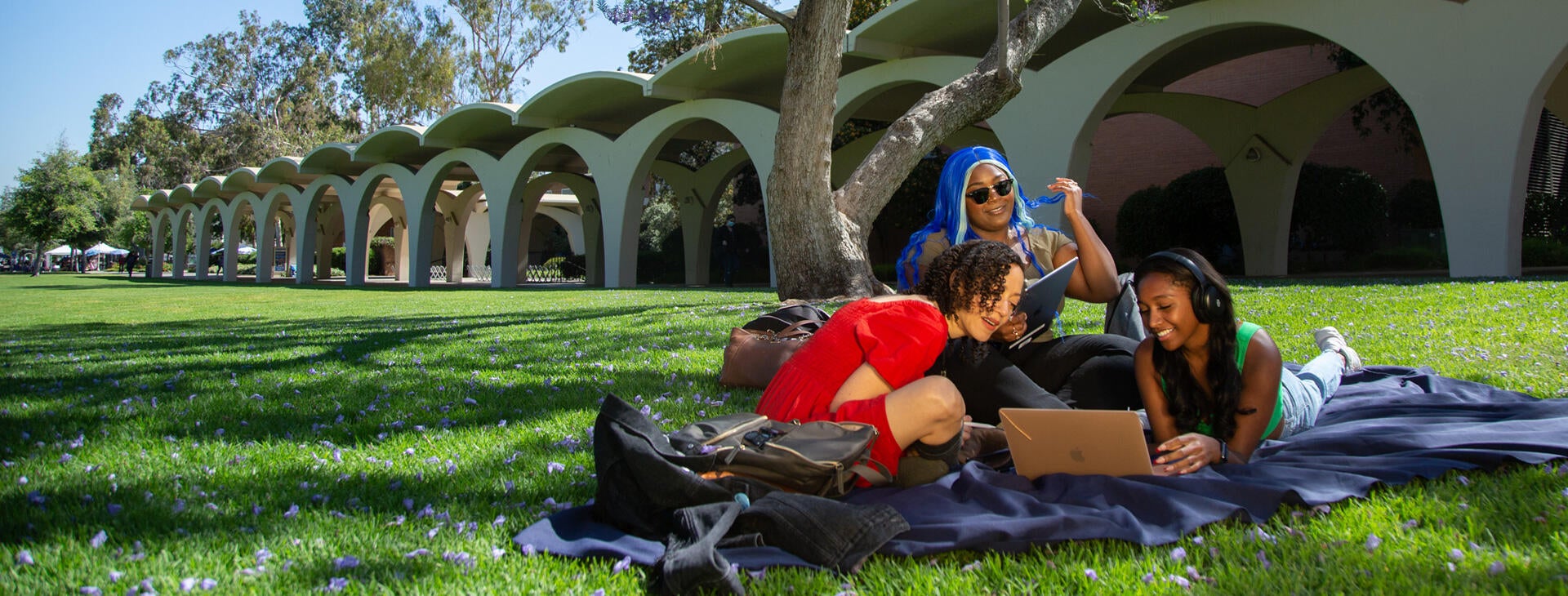 A trio of UCR students gather and sit on a blanket on the Rivera Library mall lawn. The students are all watching a laptop together and enjoying the nice day. The arches of the Rivera library are behind the students and the ground they sit on is a lush and vibrant green.