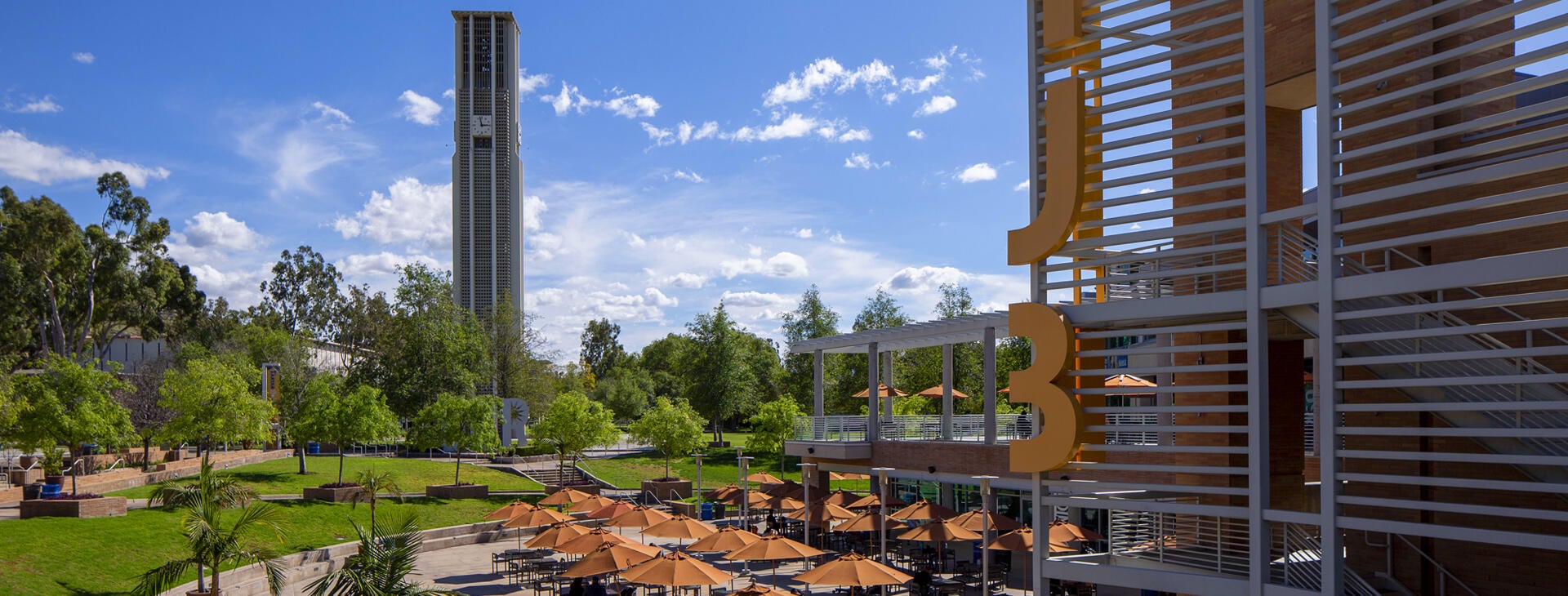 The UCR Bell Tower on a sunny day, overlooking an outdoor plaza with students studying under many orange umbrellas.