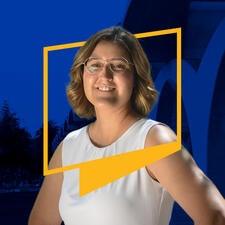 Alumna Ginny Winters smiles while wearing eyeglasses, wavy blonde-brown hair, and a white top. Her upper body is framed by a UCR icon border. The blue-tinted background features the iconic Tomás Rivera Library arches on campus.