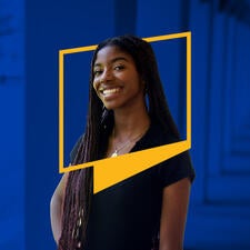 Alumna Maeve-Darly Domond smiles while wearing long, dark black hair, and a black top. Her upper body is framed by a UCR icon border. The blue-tinted background features the iconic Tomás Rivera Library arches on campus.