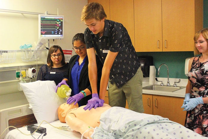 UCR_SOM_Student_Pipeline_Programs In a ER-syled hospital room, a man practices chest compressions on a CPR dummy while a woman provides the dummy with breathing assistance. Two other women observe the action.