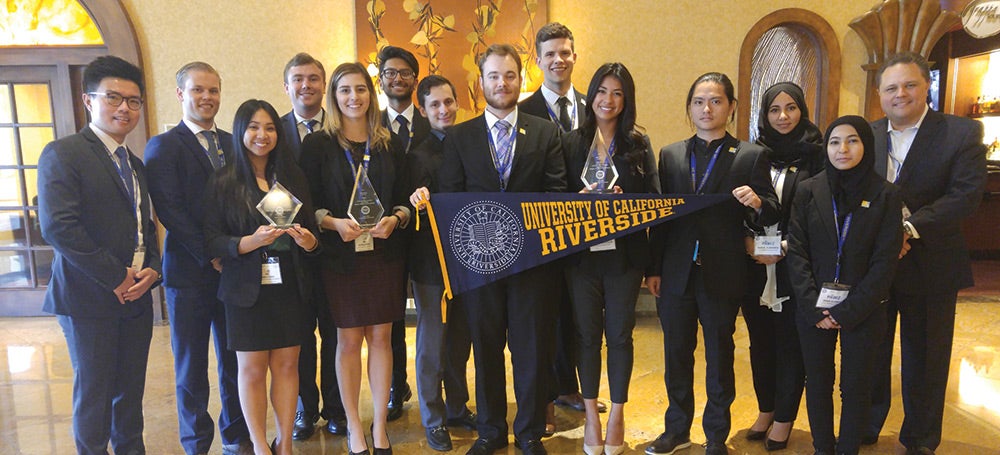 The members of the UCR Business Team, dressed in suits. In the front two women each hold awards, while another man and woman hold a UCR pennant.