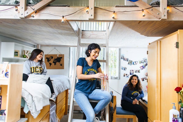 Three UCR students hanging out inside a dorm room, while wearing UCR-branded clothing.