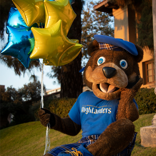 UCR's mascot, Scotty the bear, poses in front of the Business School building while wearing typical UCR tartan regalia and holding gold and blue start-shaped balloons. 