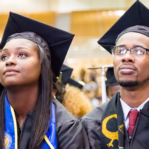 Female and male black students wearing a black cap and gown stare off during their UCR graduation ceremony. 